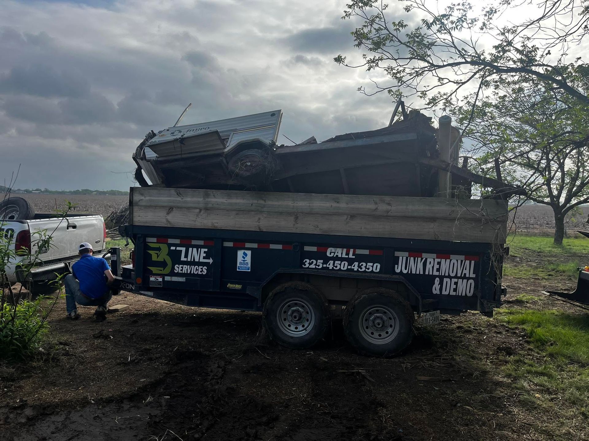 Two men load a blue dump trailer with scrap metal under a cloudy sky.