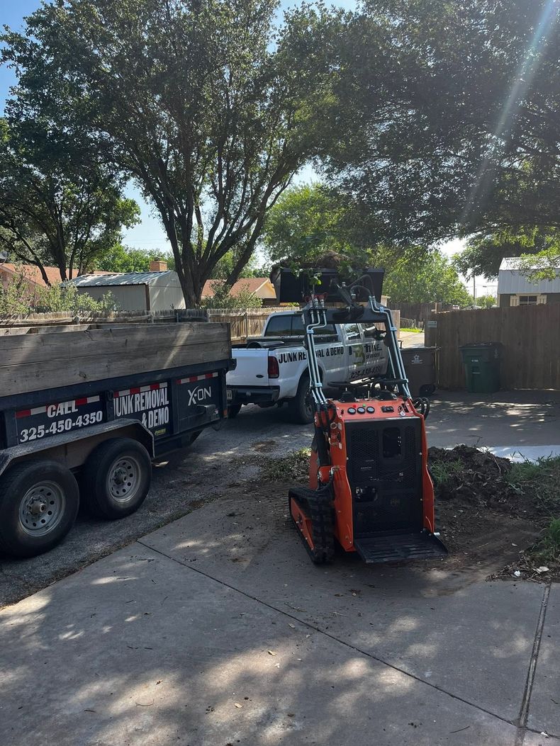 Orange skid steer loading dirt into a black dump trailer in a sunny driveway, with a white pickup nearby.