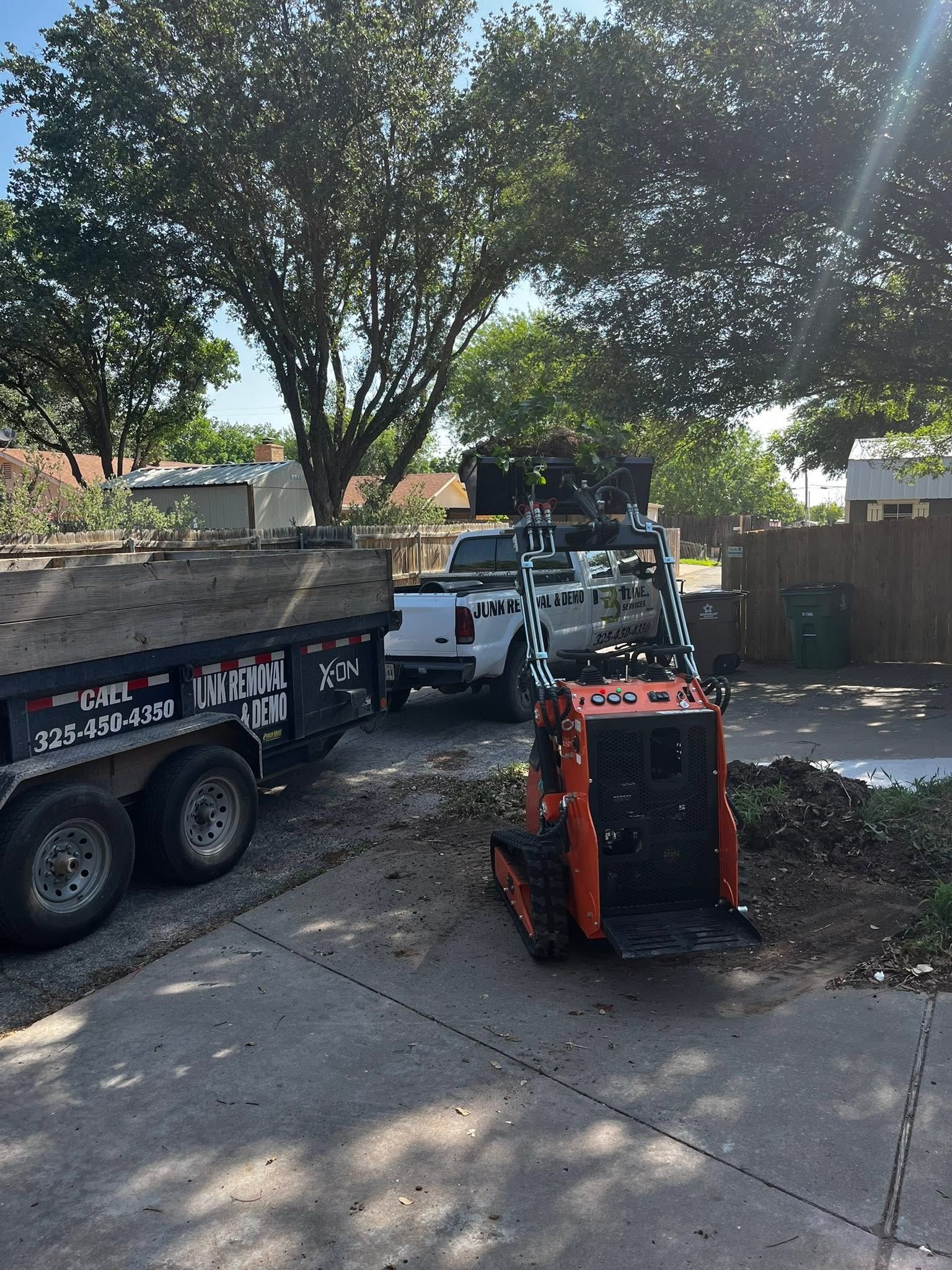 Orange skid steer beside a dump trailer and truck in a sunny yard with trees and gravel
