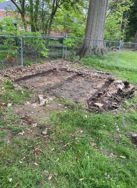 Cleared garden bed beside a chain-link fence and large tree, with soil and debris on the ground