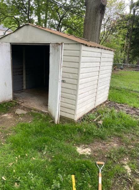 Open white backyard shed with a dark interior, grass around it, and a shovel in the foreground.