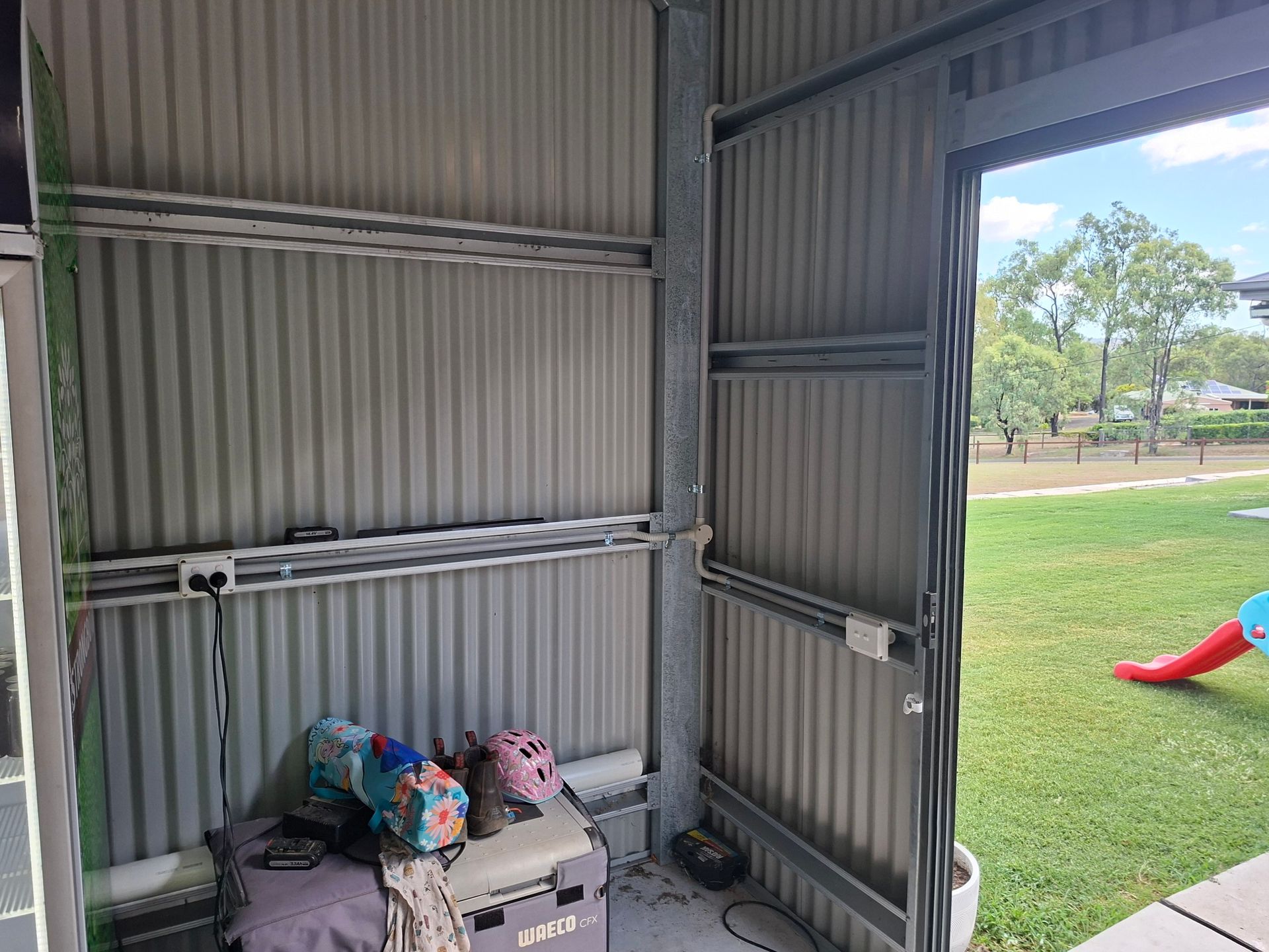 Inside a corrugated metal shed with stored items, open doorway showing a grassy park and slide outside.