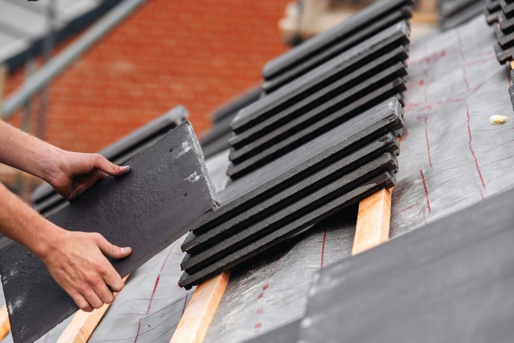 A close-up view of hands placing a dark roof tile onto a wooden batten structure on a residential roof.