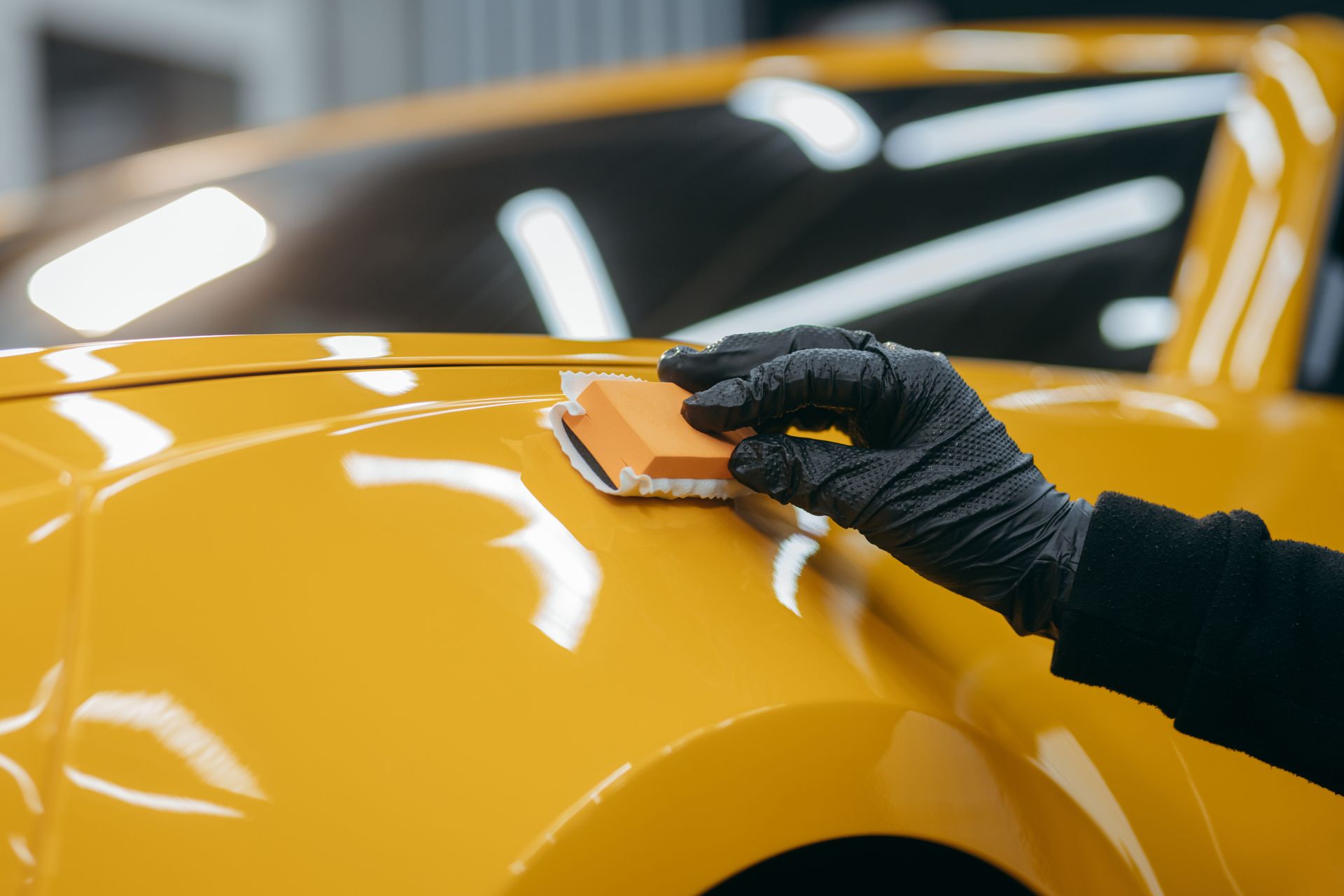 Gloved hand polishing a yellow car hood with an orange sponge