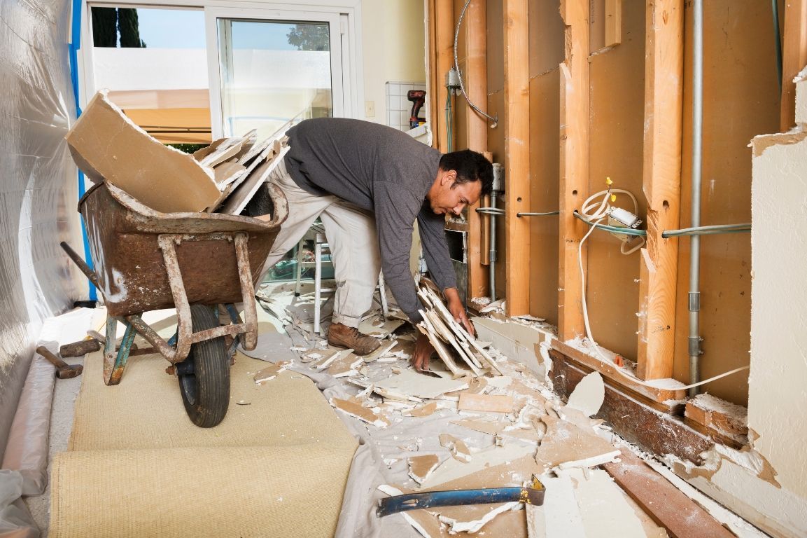 A man is kneeling down in a room with a wheelbarrow.