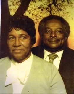 Two people in formal attire posing outdoors at sunset beneath tree branches.