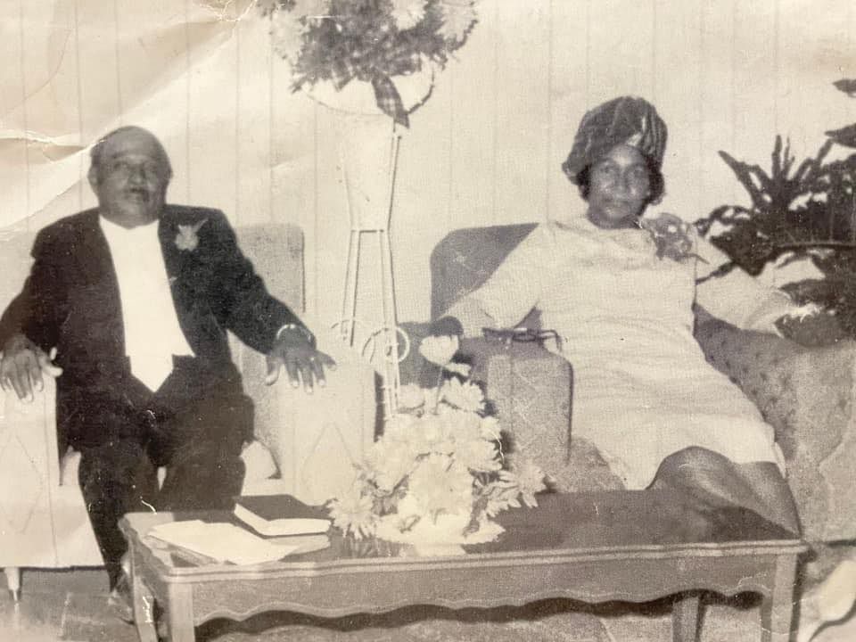 Two people seated on a couch in a living room, with a coffee table and plants nearby.