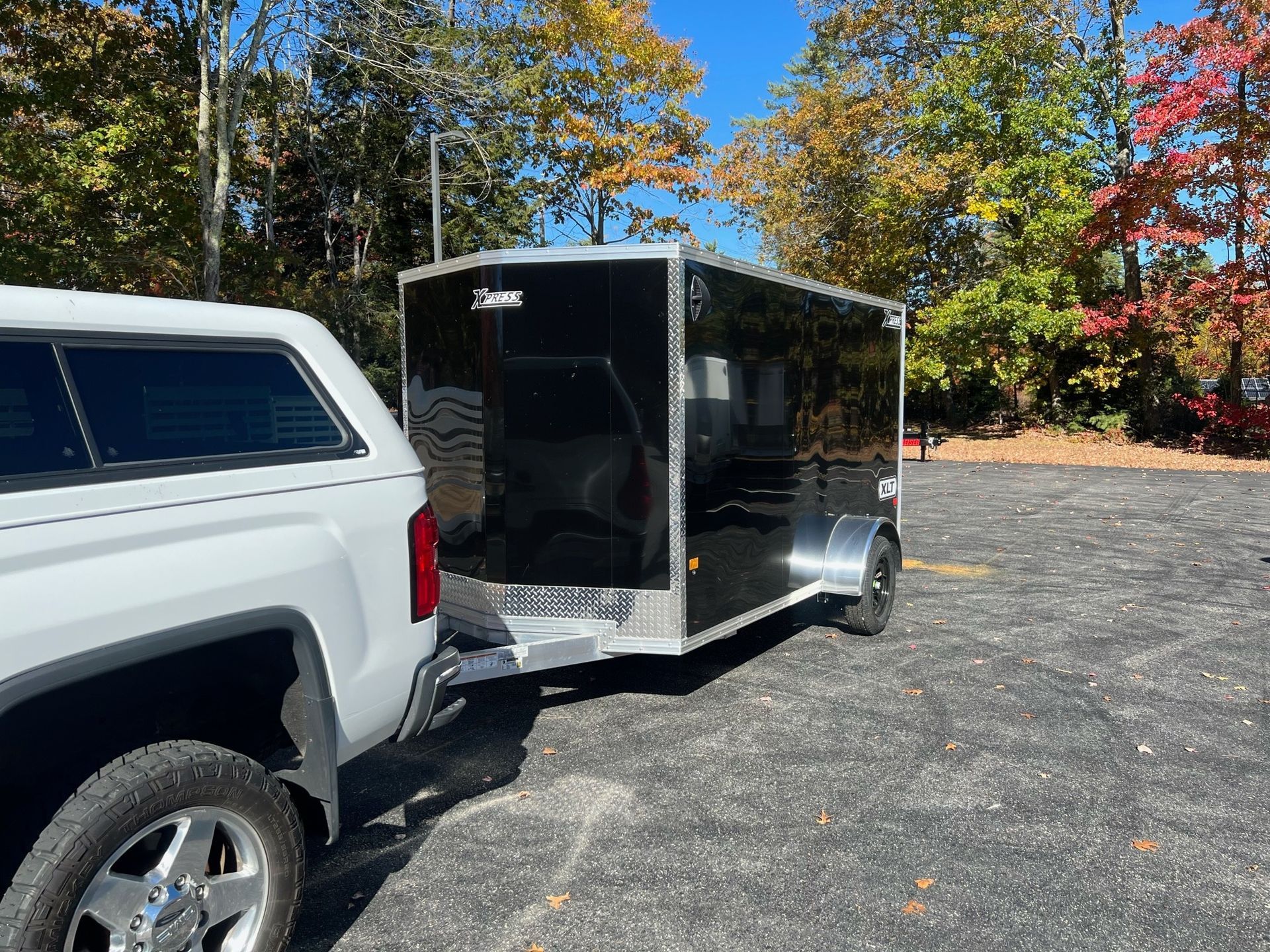 White pickup truck towing a black enclosed trailer in a gravel lot with autumn trees.