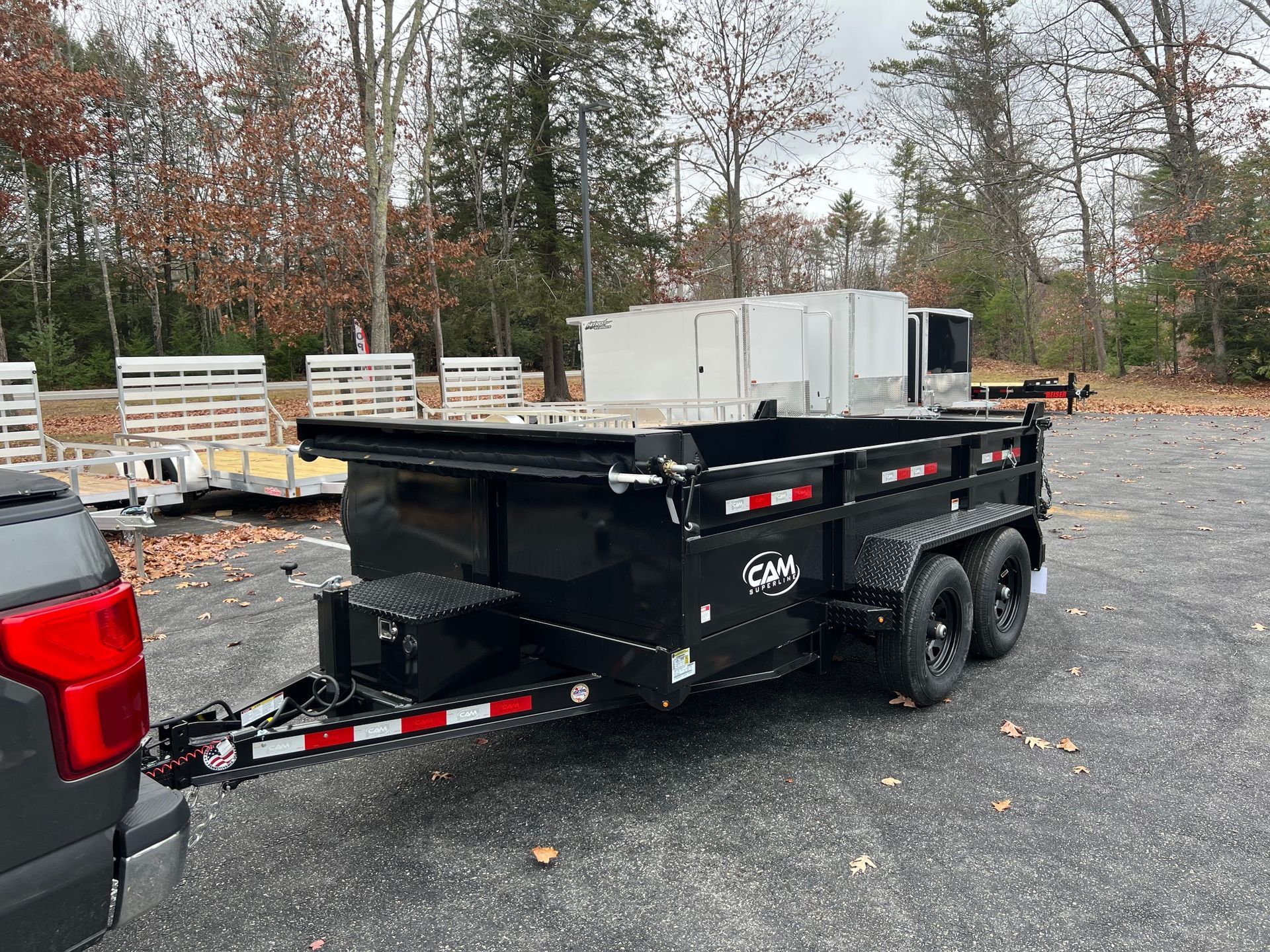 Black dump trailer with dual axles parked on gravel lot beside a pickup truck, with white equipment in back.
