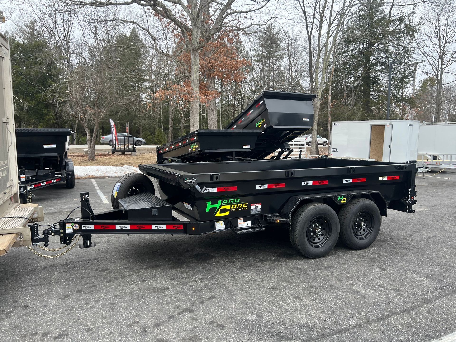 Black dump trailer with green logo parked on gravel lot, trees and equipment in the background.