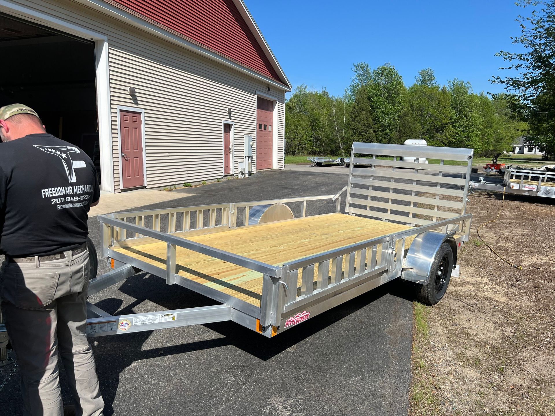 Small silver utility trailer with wooden floor parked beside a building on a sunny day