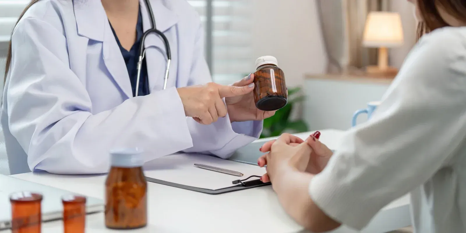 A doctor in a white coat holds a brown pill bottle and explains medication to a patient.