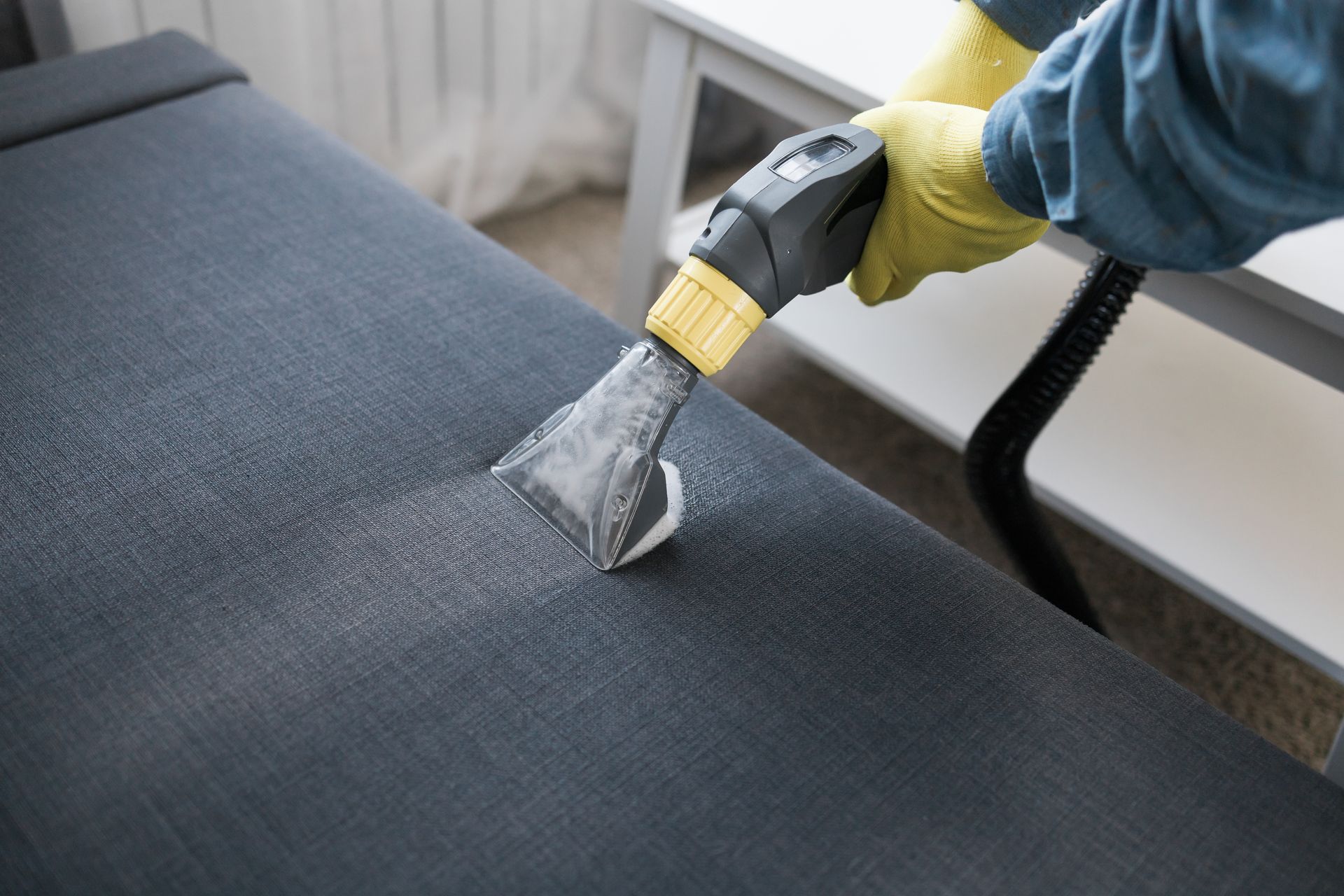 Person in yellow gloves cleaning a dark-colored upholstered surface with a steam cleaner.