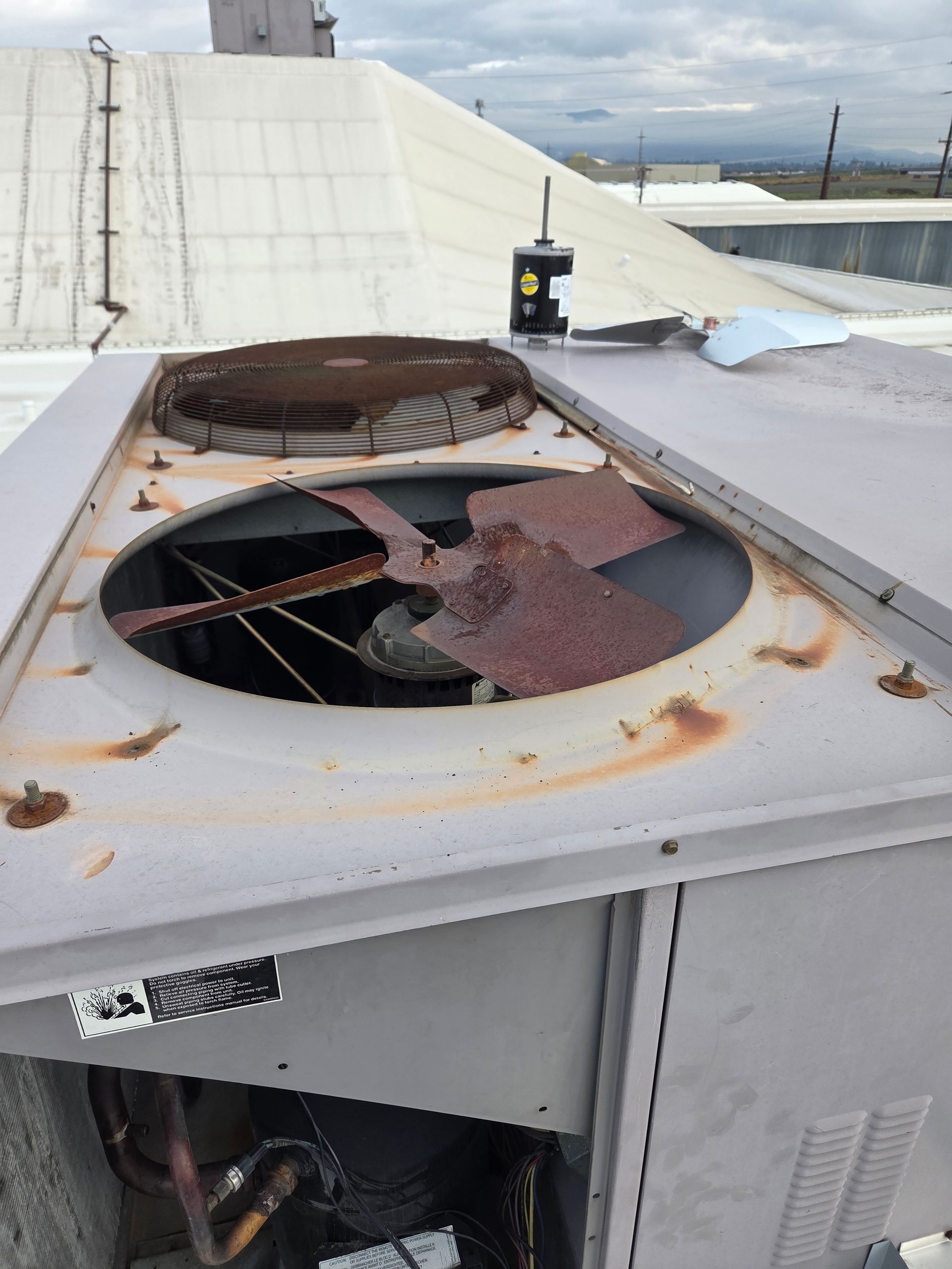 Rusted HVAC unit on a roof, with a damaged fan and exposed components. The sky is overcast.