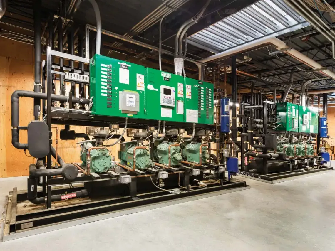 Two industrial refrigeration units with green panels, pipes, and compressors in a mechanical room.