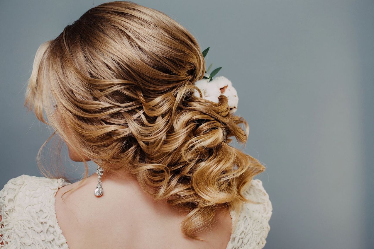 Elegante acconciatura raccolta con capelli biondi ricci e un accessorio floreale bianco su sfondo grigio.