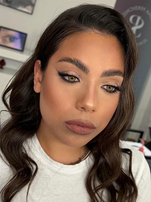 Primo piano di una persona con eyeliner alato e rossetto nude, che guarda verso il basso in una stanza illuminata da una luce soffusa.