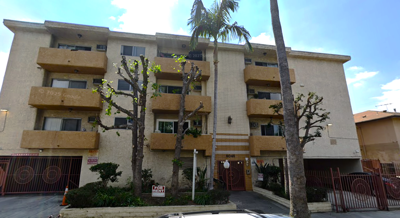 Apartment building with beige exterior, balconies, and palm trees. Brown gates on either side.