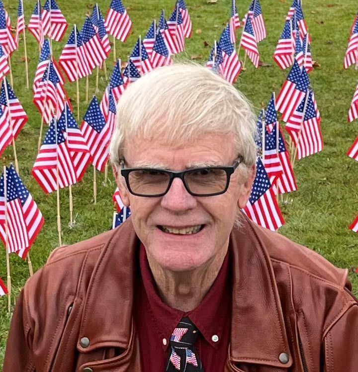 A man wearing a brown jacket is smiling in front of american flags.