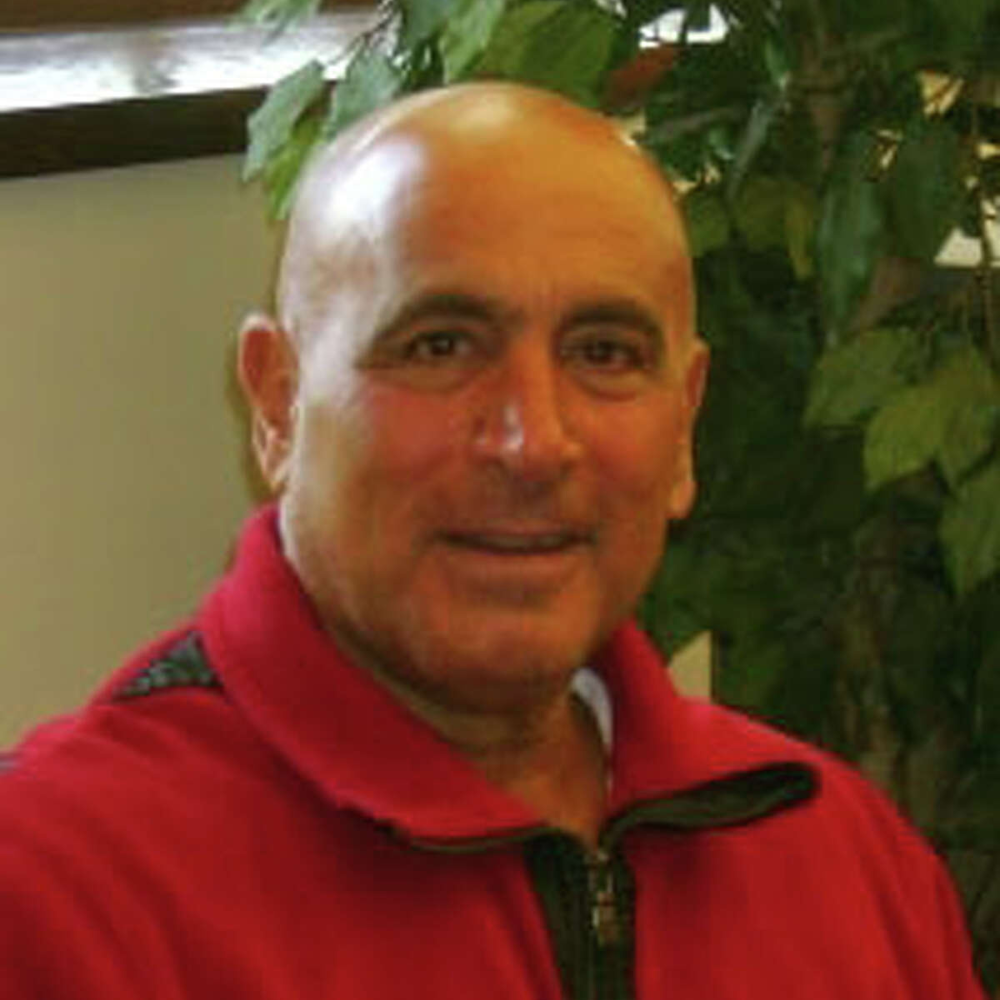 A bald man wearing a red jacket is smiling in front of a plant.
