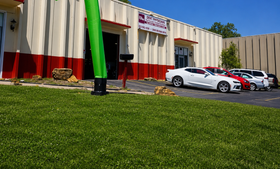 Green inflatable tube man outside a beige building with parked cars under a blue sky.