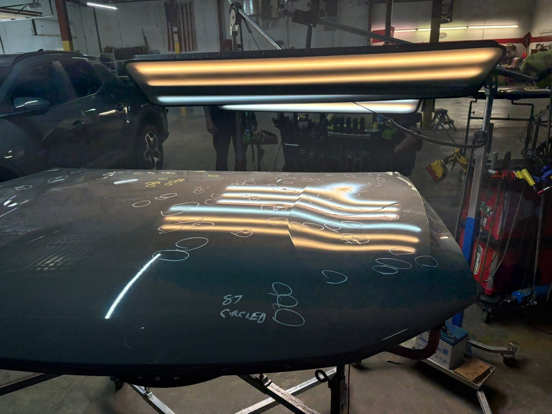Car hood in auto body shop under bright fluorescent lights, with reflections and chalk markings on the paint