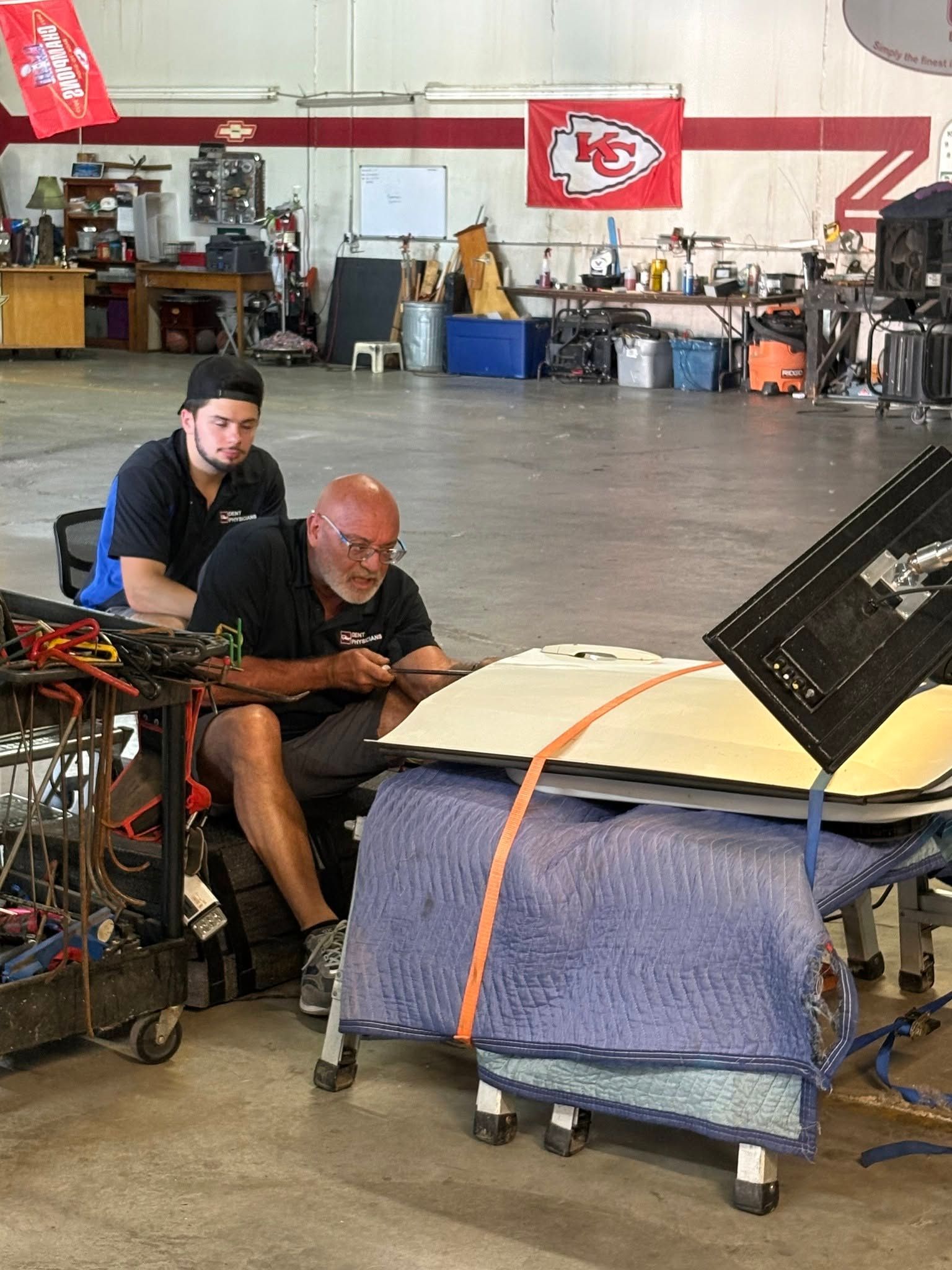 Two men crouch beside a large black vehicle panel in a workshop