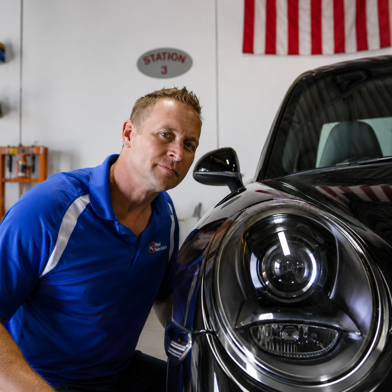 Man in blue shirt leaning beside a black car in a garage, next to a headlight.