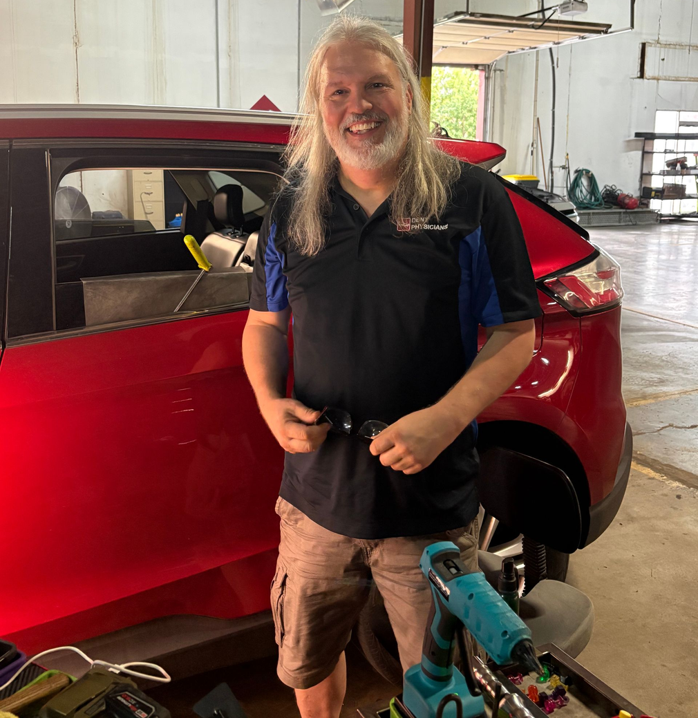 Person smiling beside a red car in a garage, holding a tool near a teal bicycle.