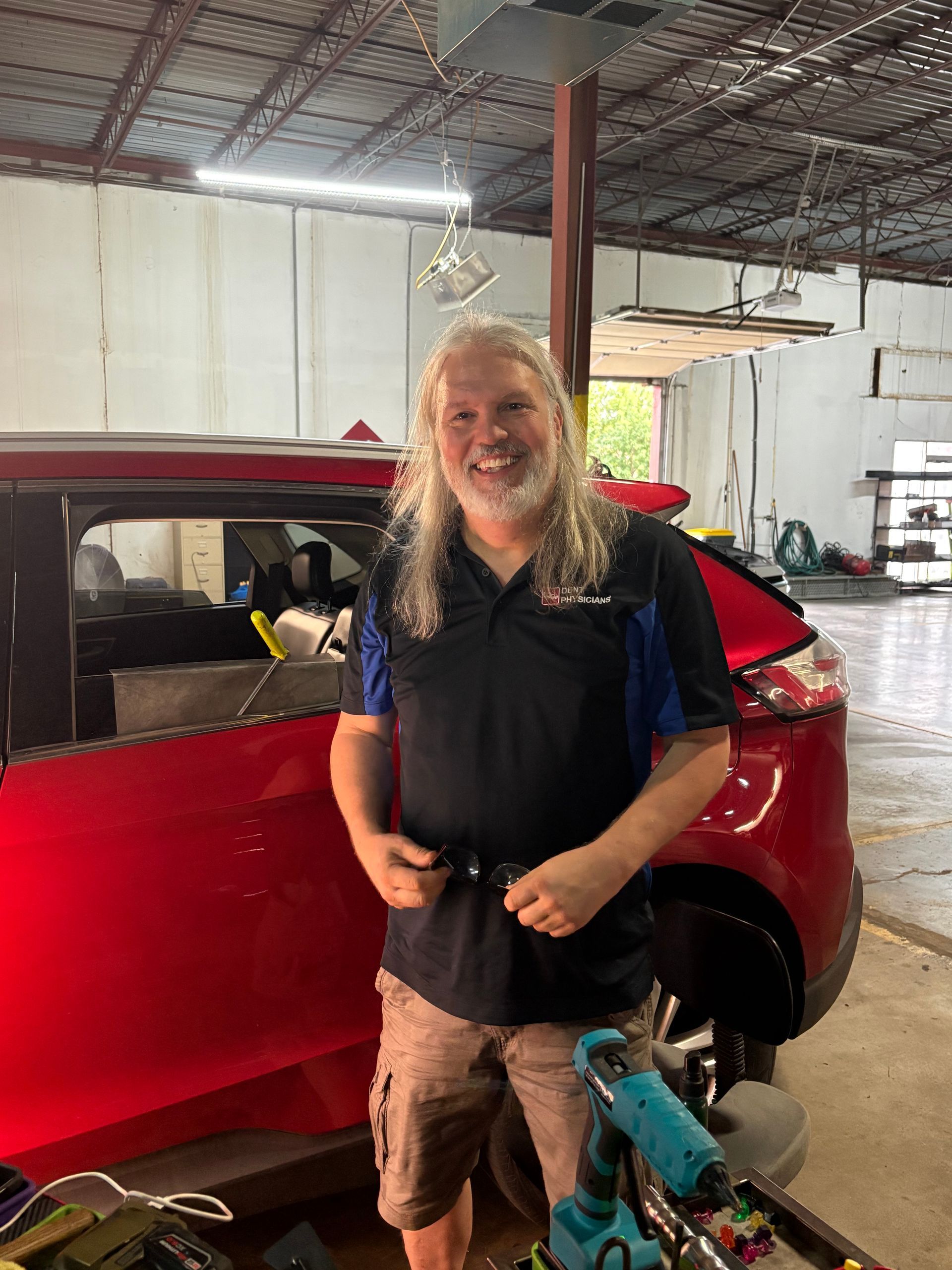 Mechanic smiling in a garage beside a black car with tools and lifts in the background