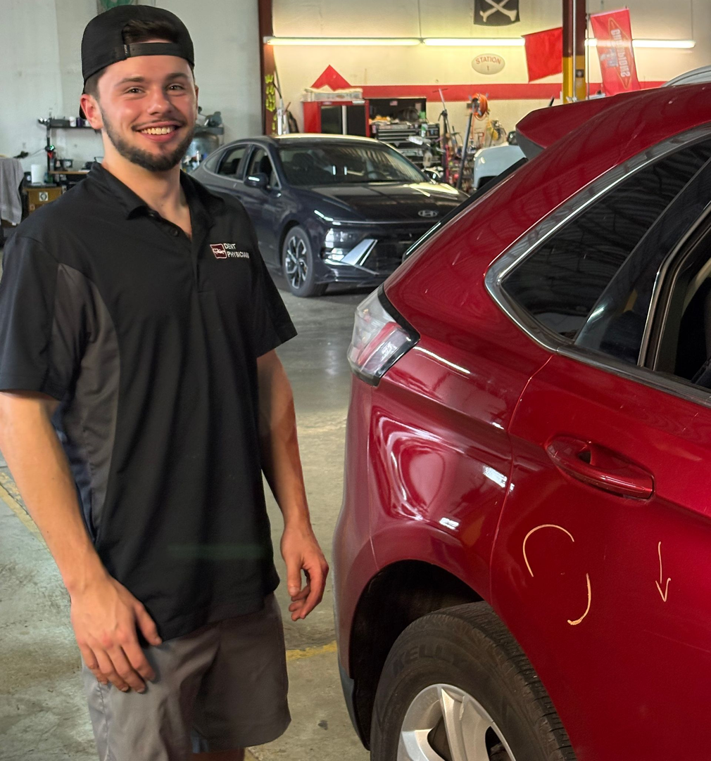 Smiling man in a black shirt standing beside a red SUV in a car showroom
