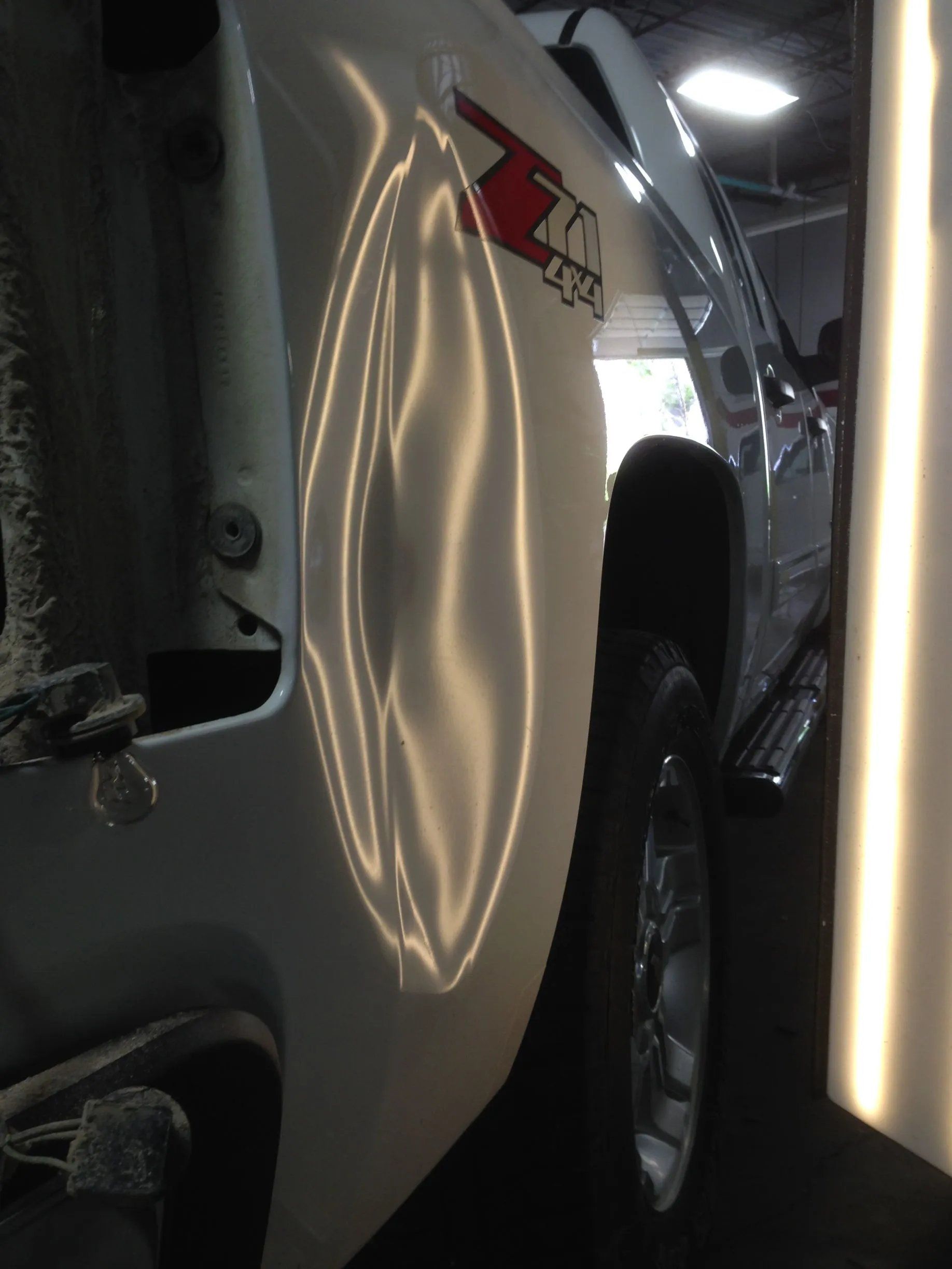 Silver SUV in a dim garage, close-up of side panel with a large dent and a vertical light reflection