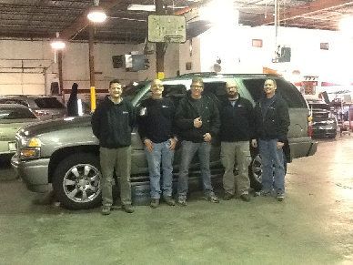 Five people posing beside a silver SUV in a garage, under bright overhead lights.