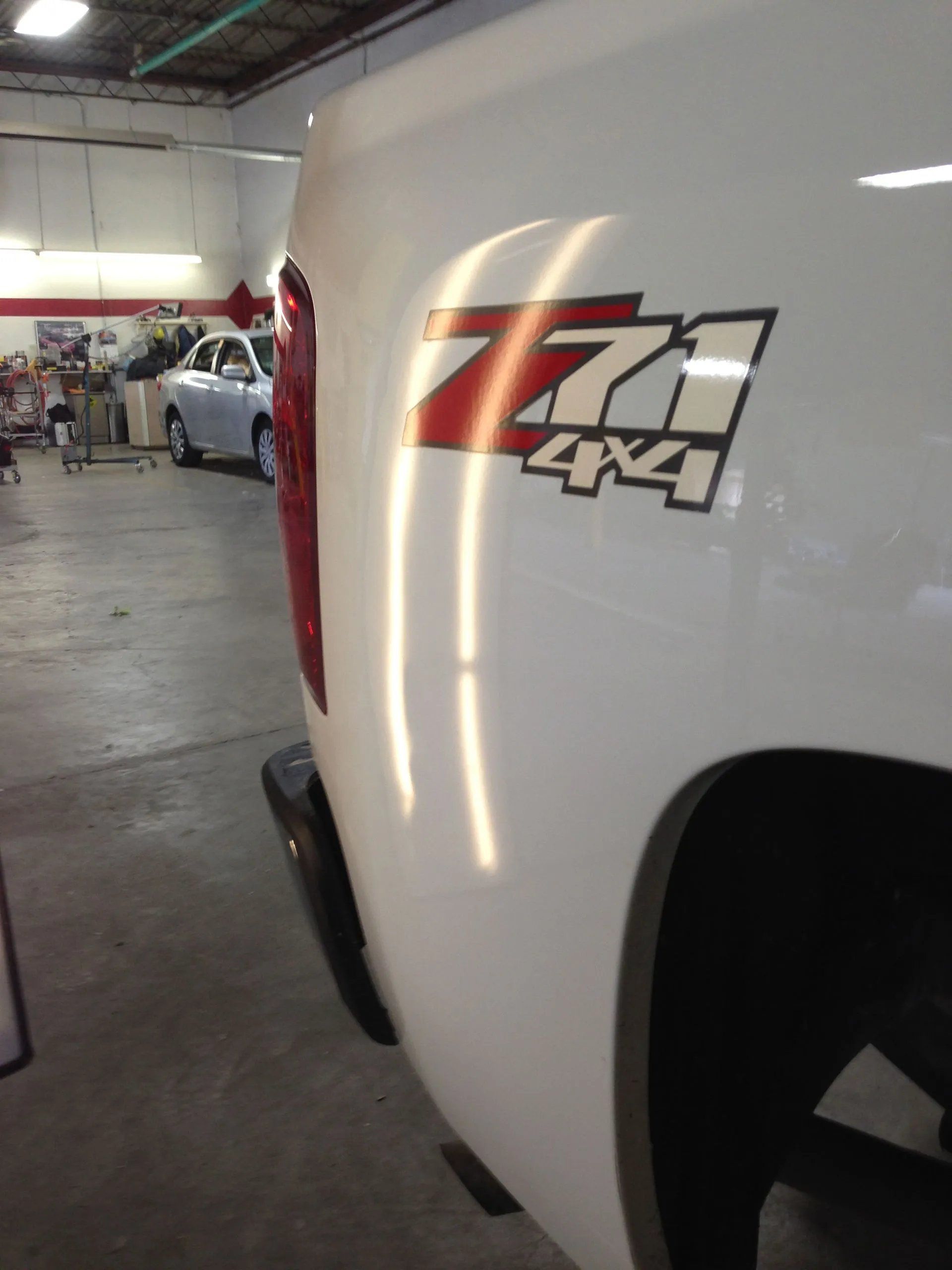 Close-up of a white Chevy 4x4 truck with a red stripe in a garage, showing the rear side panel.