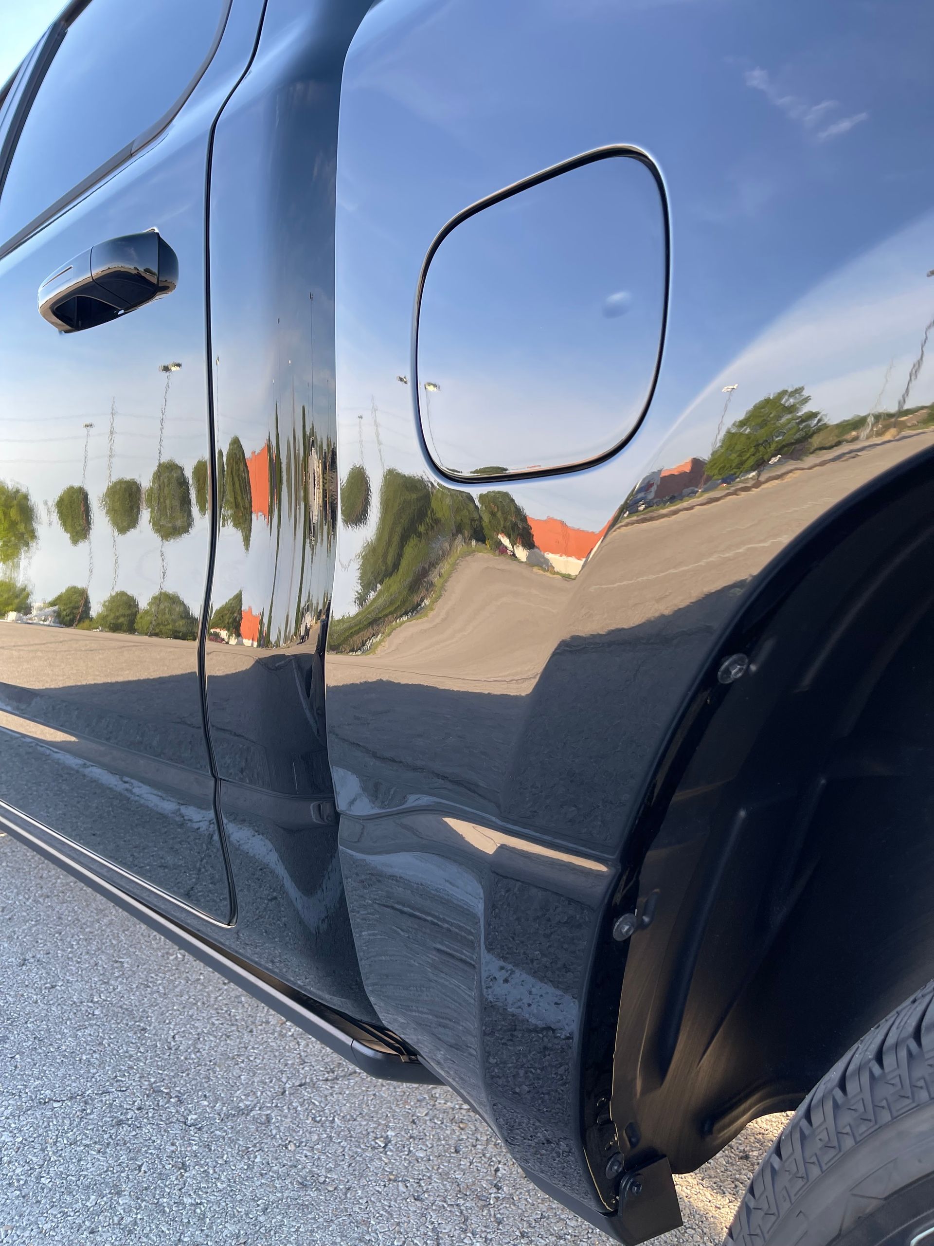 Close-up of a glossy black car side panel with fuel door, reflecting the road and trees.