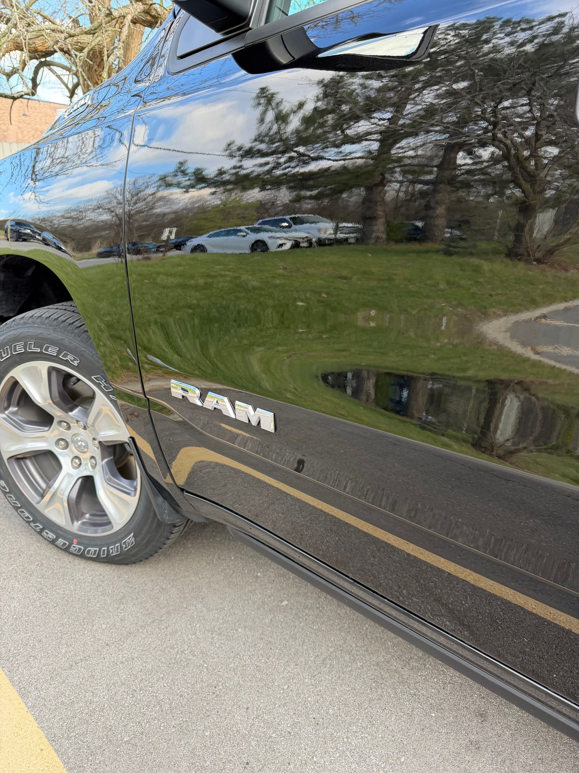 Close-up of a black RAM truck door and front wheel reflecting trees and a road.