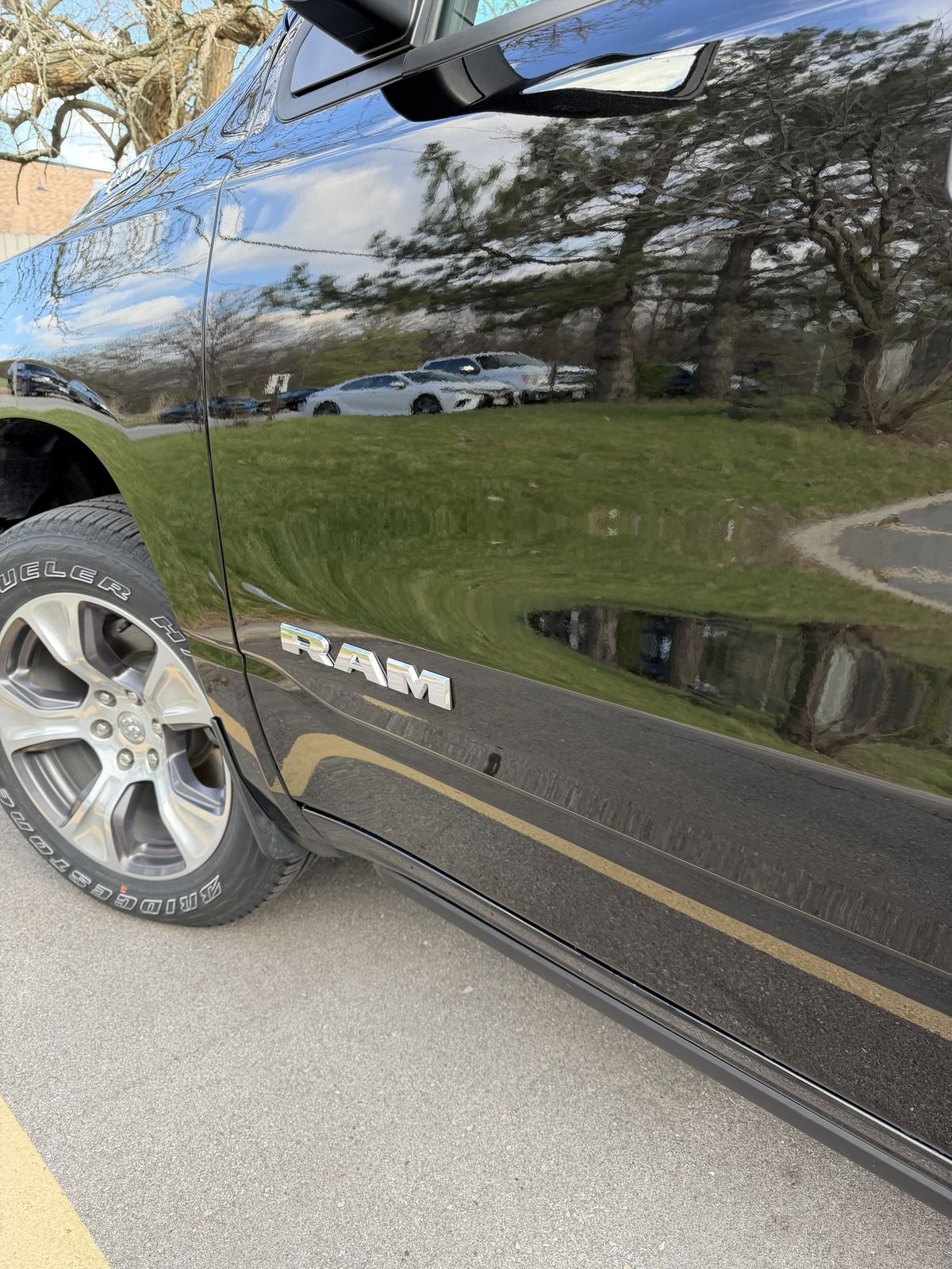 Black RAM truck parked roadside, showing glossy side panel and front wheel reflected with trees and sky.