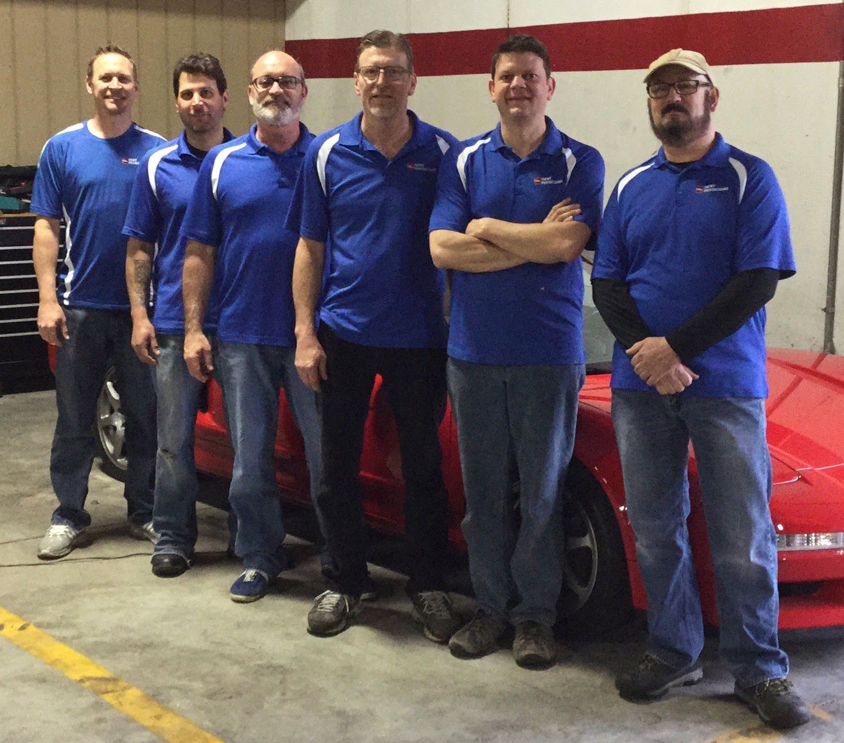 Six people in matching blue shirts stand beside a red car in a garage.
