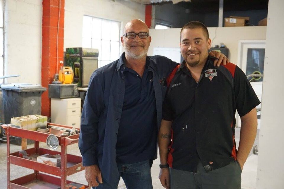 Two smiling men standing together in a workshop, one in a navy jacket and one in a black shirt.