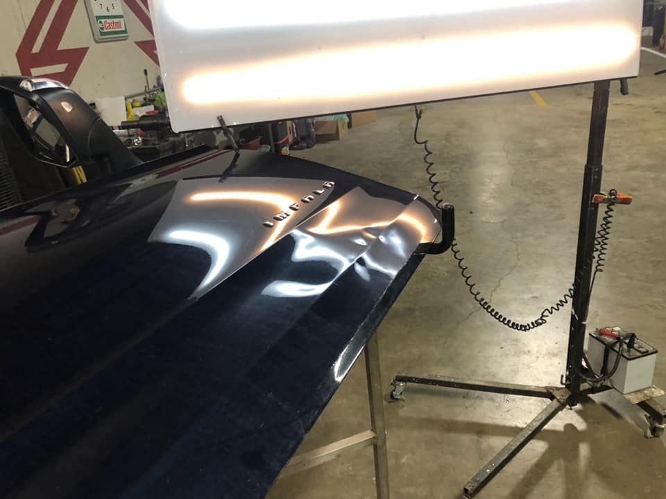 Car hood under bright shop lights, with visible dents and reflective dark blue paint.