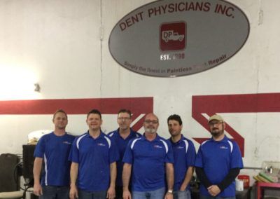 Five people in blue shirts pose in a mechanic shop beneath a DENT PHYSICIANS INC. sign