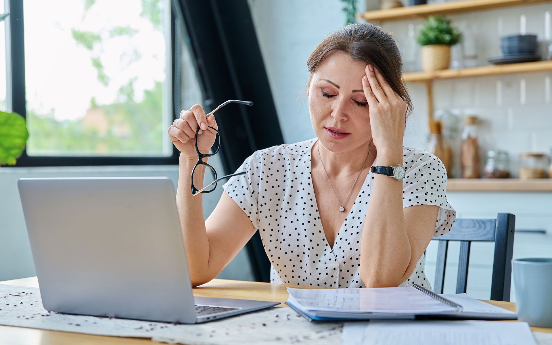 Woman with headache sits at desk with laptop, holding glasses and touching forehead.