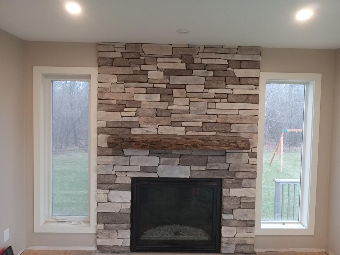 Stone fireplace with wooden mantel between two tall windows in a bright room