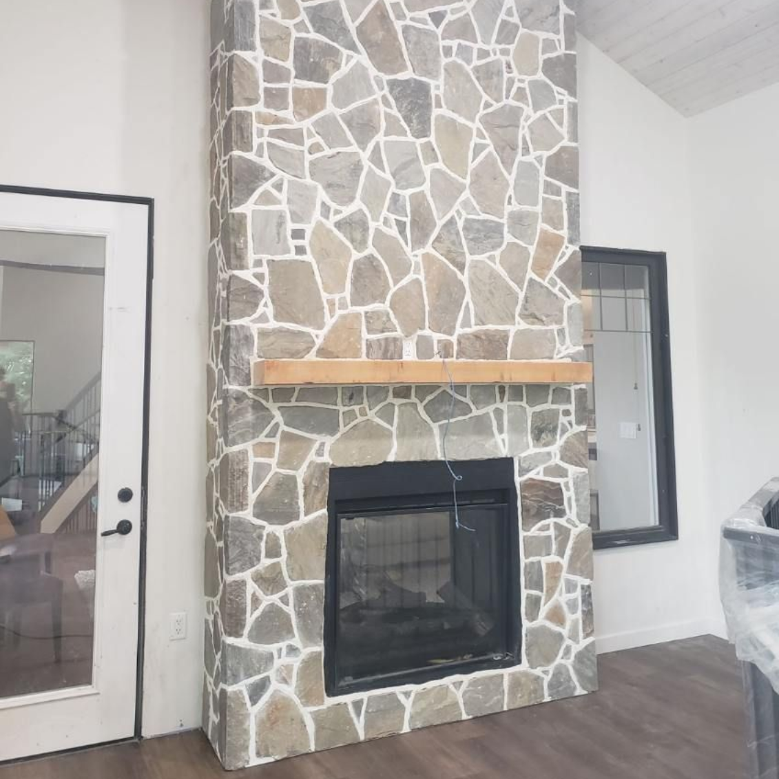 Stone fireplace with wood mantel in a bright room, next to a glass door and window.