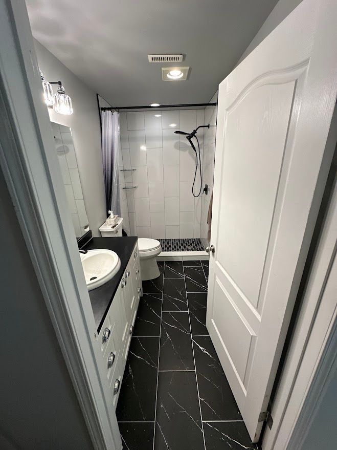 Narrow gray bathroom with black tile floor, white vanity, sink, shower, and white door.
