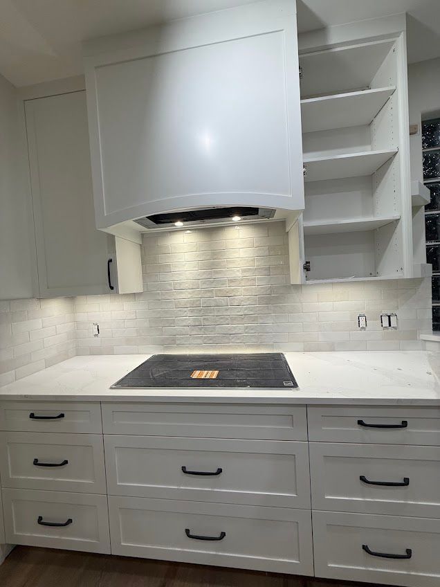 Modern white kitchen with built-in cooktop, stainless hood, gray backsplash, and open shelving.