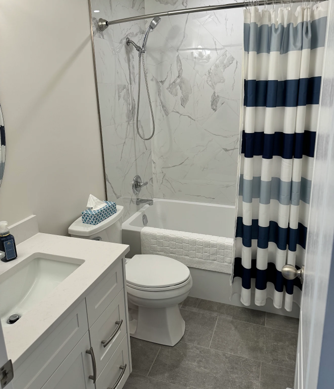 Small modern bathroom with gray tile, white vanity, toilet, and striped shower curtain over tub.
