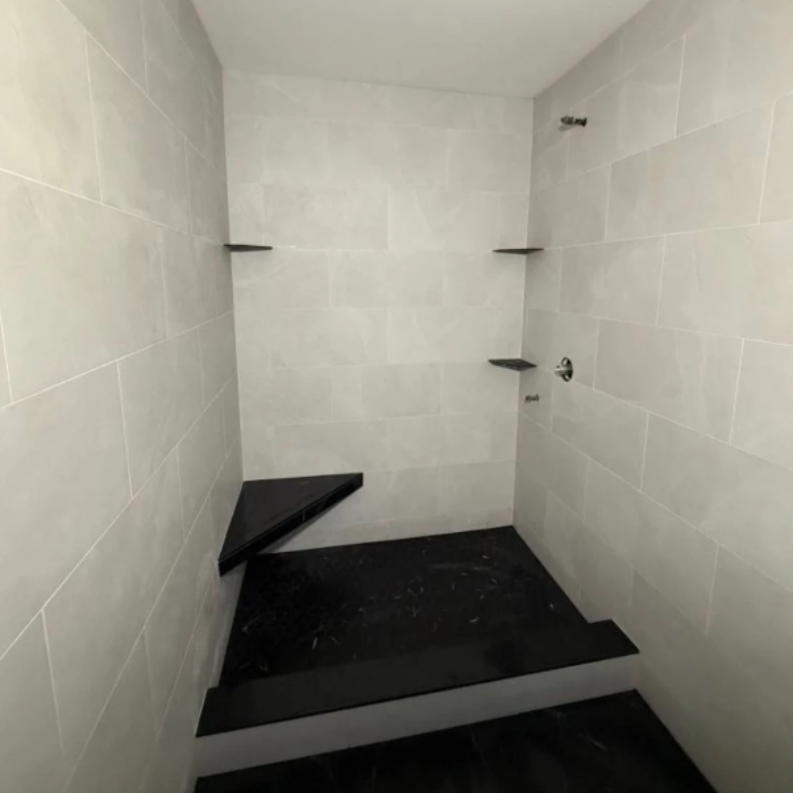 Empty tiled shower stall with black floor and built-in corner bench in progress