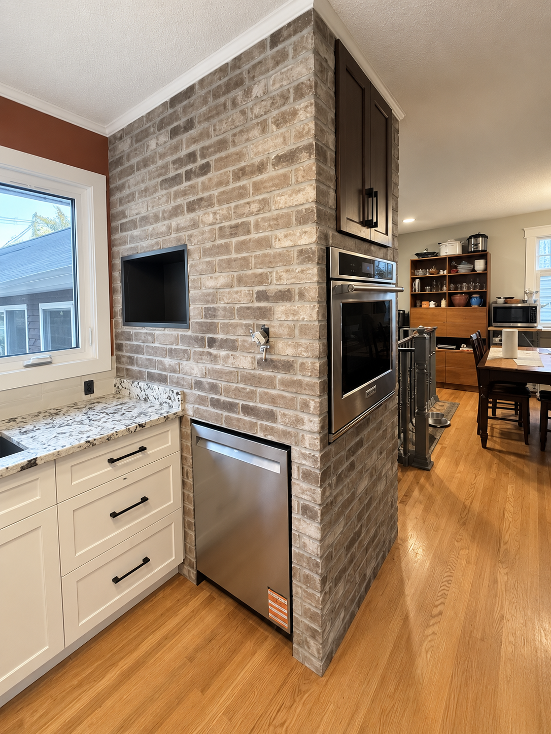 Kitchen corner with brick column, built-in oven, stainless dishwasher, and window above countertop