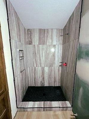 Newly tiled shower stall with gray and white wood-look tiles, black hexagon floor, recessed shelf.
